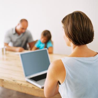 Woman working on laptop with husband and child in background - The Juggling Act