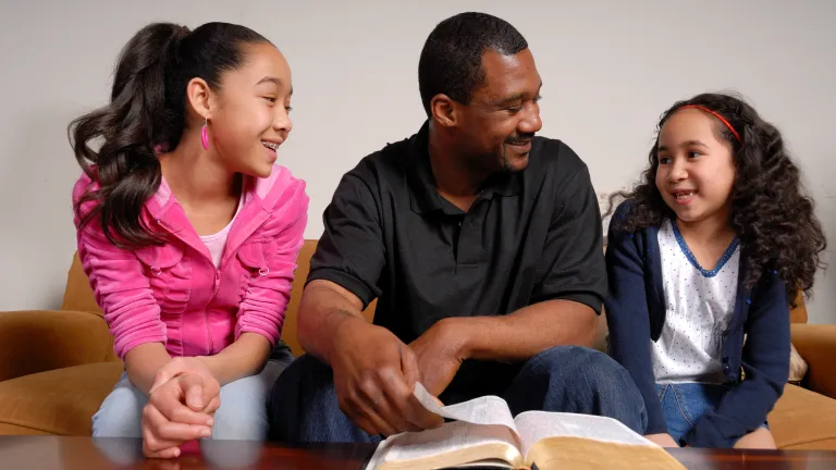 As we navigate the challenges of a world sliding away from God toward the eager embrace of the devil, whose influence is both subtle at times and overt at others, our families can be a bastion of righteousness. A dad and his two daughters talking with a Bible opened.
