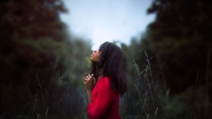 a woman wearing and red dress kneeling with folded hands outdoors amidst trees