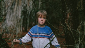 A little boy standing by trees in a forest.