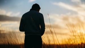 A man looking down while standing in a field. The sun is setting in the distance.