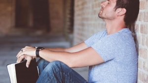 A man holding his Bible with his back propped against a wall - with eyes close and head down praying.