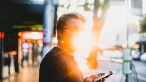 A man holding a smartphone with the sun glaring behind him.