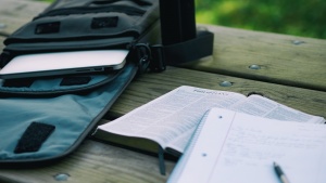 A Bible and notebook laying on a table. A laptop is in backpack.