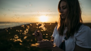 A woman holding a dandelion with the seeds of the plant blowing away.