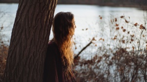 A young woman leaning against a tree looking out over a lake.