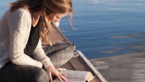 A woman sitting on a dock in the water reading her Bible.