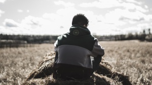 A young man sitting on a round bale of hale looking out over a field. 