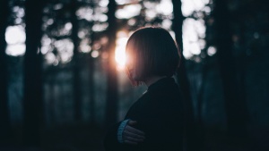 A young woman standing in a patch of trees with the sun shining through.