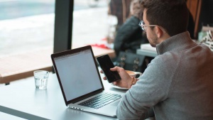 A man using his laptop and smartphone while sitting at a table in a cafe.