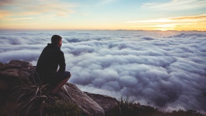 Photo of man thoughtfully squatting at the top of a cliff looking at the clouds.