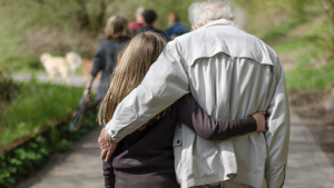 Photo of a young girl on the left and old man on the right, walking along a paved trail in a park, each with an arm around the other.