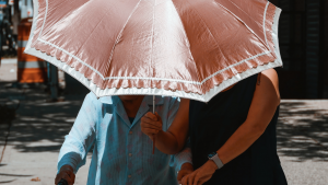 Photo of two women walking under a parasol; an elderly woman with a walker being helped by a middle-aged woman.