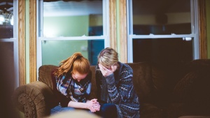 Photo of two women sitting on the couch grieving together. One woman has her hands clasped and one woman is leaning her face into her forhead.