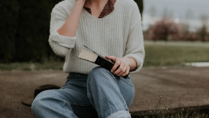 A girl in cropped mom jeans sits on the ground with a Bible in her lap. A wide brim hat sits next to her and she has a flowery collared shirt on under a white sweater.