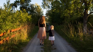 Photo of a mother and young daughter walking down a dirt road in the countryside; the daughter is holding her mother's hand and looking up at her.