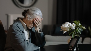 a woman with her head in her hand as she sits at a table across from white flowers and a photo frame