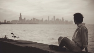 a man sitting cross-legged in a concrete ledge overlooking a body of water an a distant city skyline