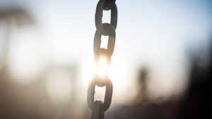 a length of chain in the foreground with the sunset shining in the background, the light hitting the chain in the center, interrupting the chain's solid appearance