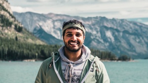 a man stands outdoors with mountains and a lake behind him