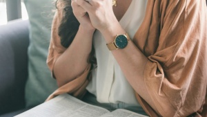a woman sitting with hands folded in prayer with an open Bible in her lap