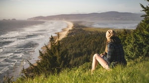 a woman sitting in the grass and looking towards the ocean shore