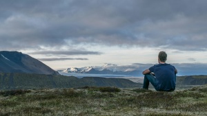 A young man sitting on the grass looking at a view of mountains.