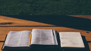 A opened Bible laying on a picnic table beside a notebook.