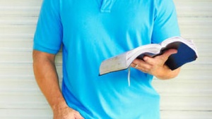 Man reading a Bible while leaning against a wall.