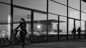 A young woman pushing a bike while walking by a office building with mirrored windows.