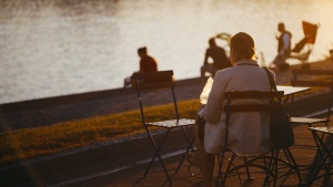 A woman sitting at table outside by a lake watching people.