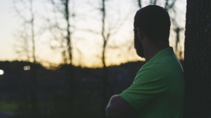 A man with arms crossed leaning against a tree looking at the sun setting.