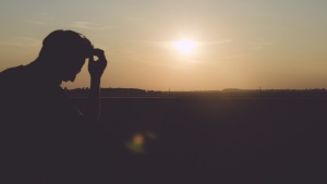 A young man sitting with head resting on tips of his finger. The sun is setting in the background.