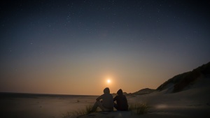 Two people sitting on a beach with the sun setting. 