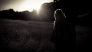 A woman standing in a field with the sun glaring behind a row of trees.