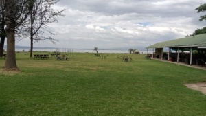picnic benches and a shelter built on an expanse of green lawn under a cloudy sky with water and mountains in the distance