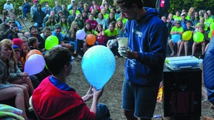 A photo of a camper holding a balloon while another camper paints it with shaving cream. The whole camp is watching in background. 