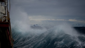 Photo of a stormy sea taken from a ship. The waves are crashing against the ship and the sky is filled with low gray clouds.