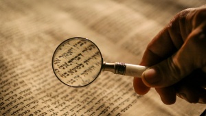 a hand holding a magnifying glass over a scroll of Hebrew text