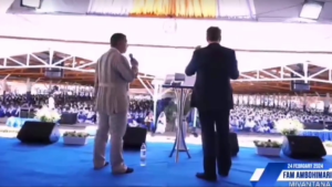 two men standing with microphones on a stage in front of a large crowd