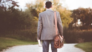 a man dressed in a suit, holding a Bible and with a satchel hanging from one shoulder, standing on a road