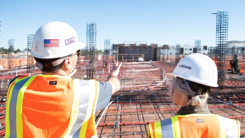 two people wearing construction hats and vests and looking over a construction site with their backs to the camera