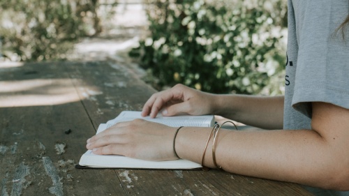 a woman's arms resting on an open Bible outdoors on a wooden table