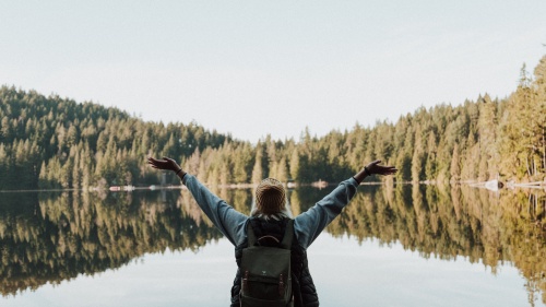 a woman with her arms raised in a joyful gesture facing a lake surrounded by trees