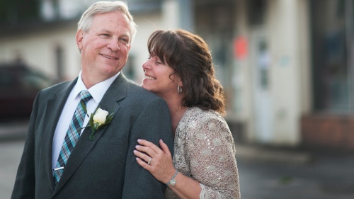 a man wearing a suit and tie and a woman wearing a dress standing behind him with a hand on his arm