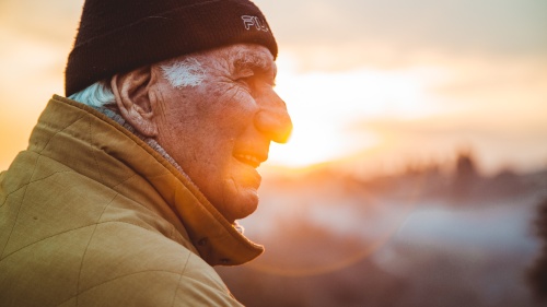 an elderly man wearing a hat and coat as he stands outdoors looking into the distance with a sunset in the background