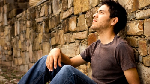 A young man sitting against a rock wall.