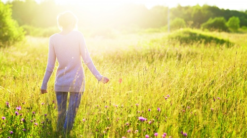 A woman walking in field of tall grass and flowers as sun rays shine on her.
