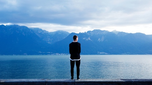 a man wearing black standing with his back to the viewer as he faces a blue mountainous landscape over a body of water