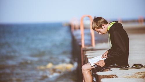 A young man reading a Bible while sitting on a dock.